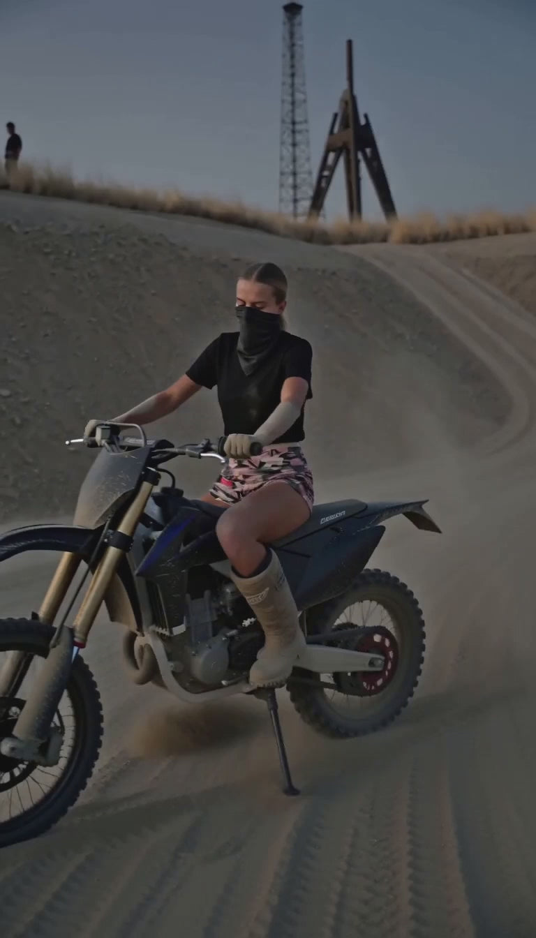 Szorty Slim Camo Nowrocky™ — osoba w chuście na twarzy prowadzi motocykl w poślizgu po wydmie, szorty moro widoczne, stalowa wieża na grzbiecie wydmy, zmierzch.
