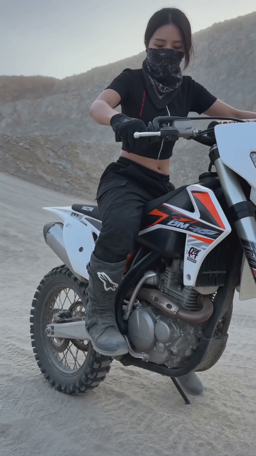 T-shirt Crop z Zamkiem Nowrocky™ — kobieta z chustą na twarzy zsiada z motocykla enduro na żwirowym zboczu, czarny top crop z zamkiem i czerwonym nadrukiem, kurz w powietrzu.