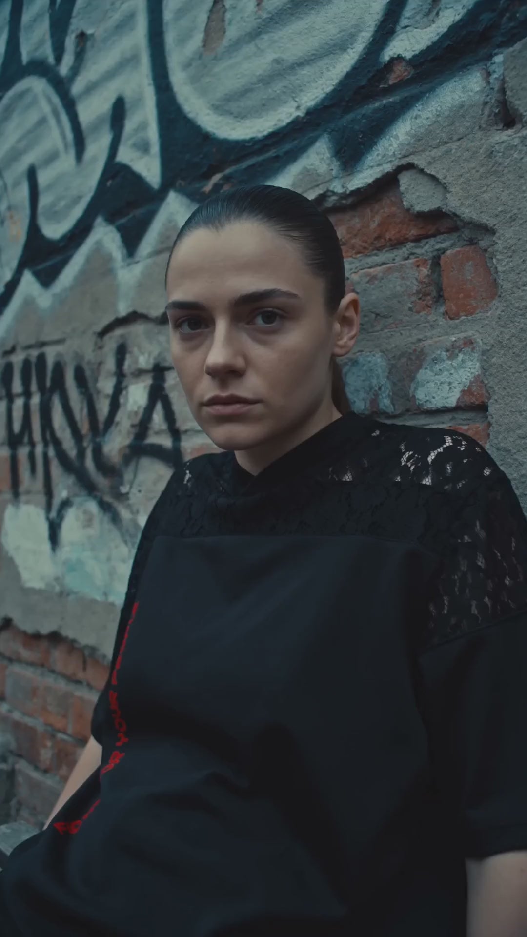 T-shirt z Koronkowym Panelem Nowrocky™ — kobieta pod ceglaną ścianą pokrytą graffiti, czarny top z koronkowym panelem na ramieniu, czerwony pasek nadruku na boku.