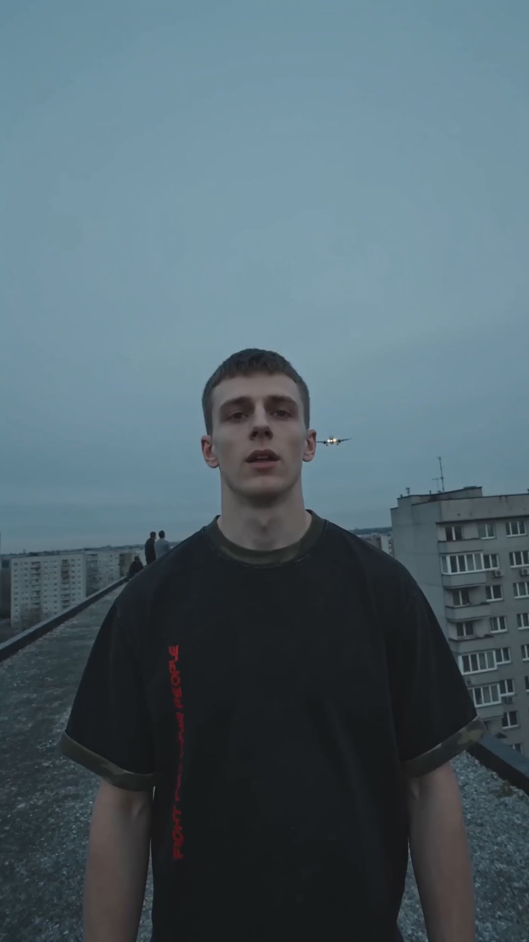 T-shirt Boxy Camo Mineral Wash Nowrocky™ — mężczyzna stoi na dachu bloku, szare niebo, w tle dachy osiedla, koszulka z czerwonym nadrukiem na boku.