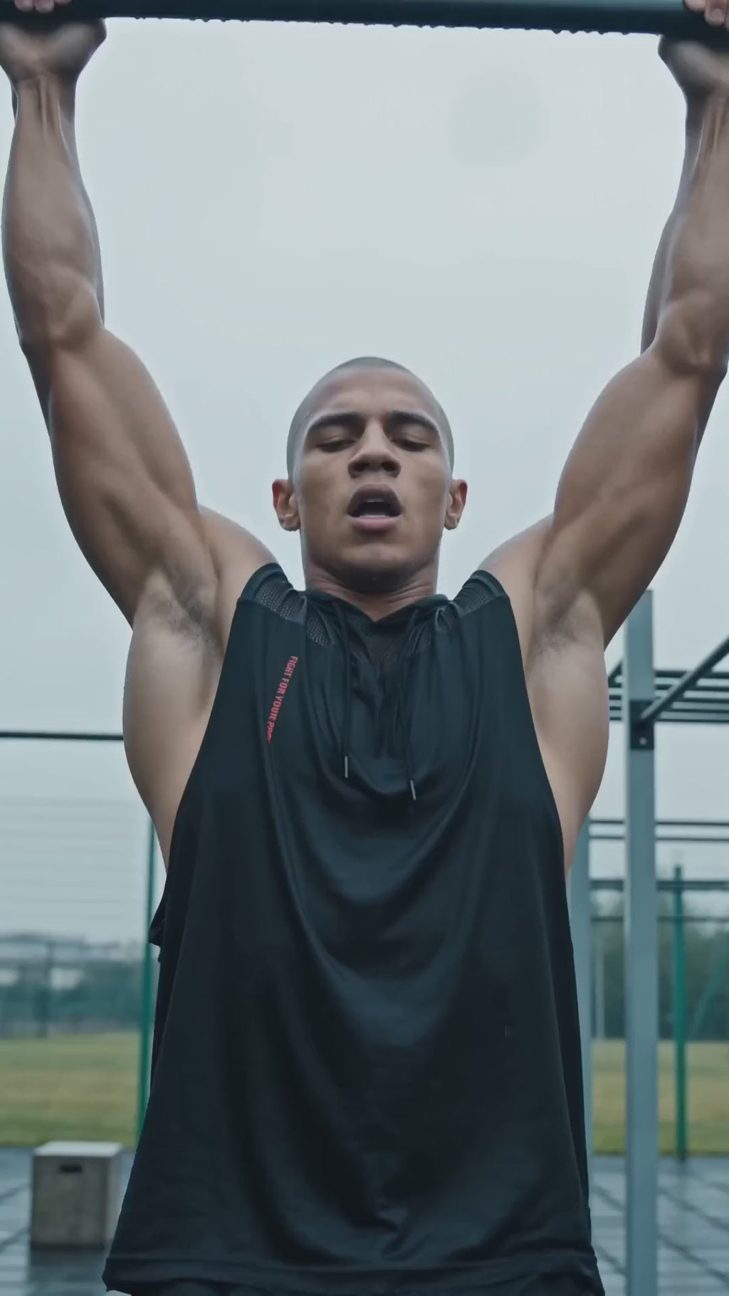 Tank Top Mesh z Kapturem Nowrocky™ — mężczyzna wisi na drążku podczas podciągania na osiedlowej siłowni pod chmurką, czarny bezrękawnik z kapturem i czerwonym nadrukiem.