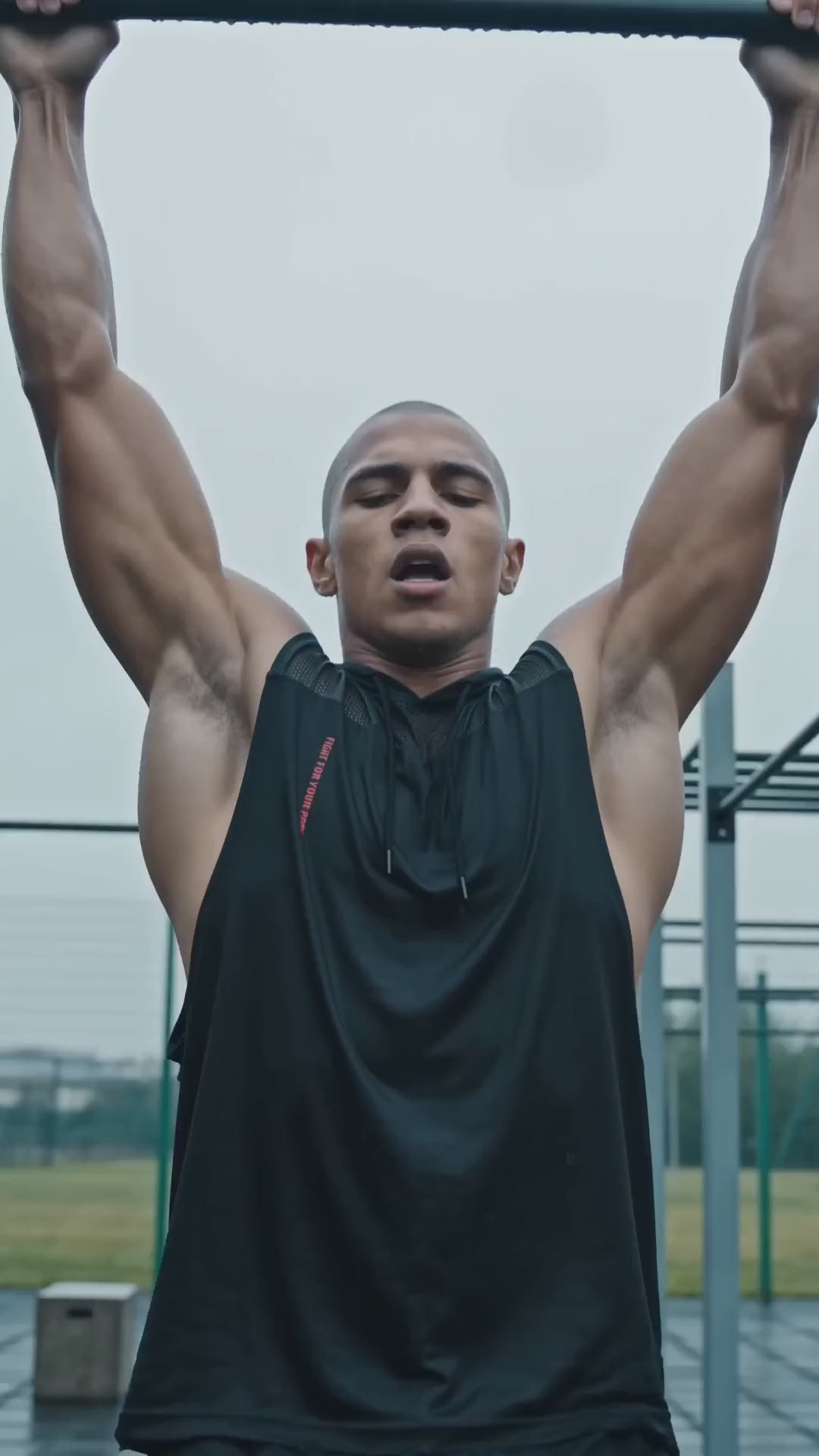 Tank Top Mesh z Kapturem Nowrocky™ — mężczyzna wisi na drążku podczas podciągania na osiedlowej siłowni pod chmurką, czarny bezrękawnik z kapturem i czerwonym nadrukiem.