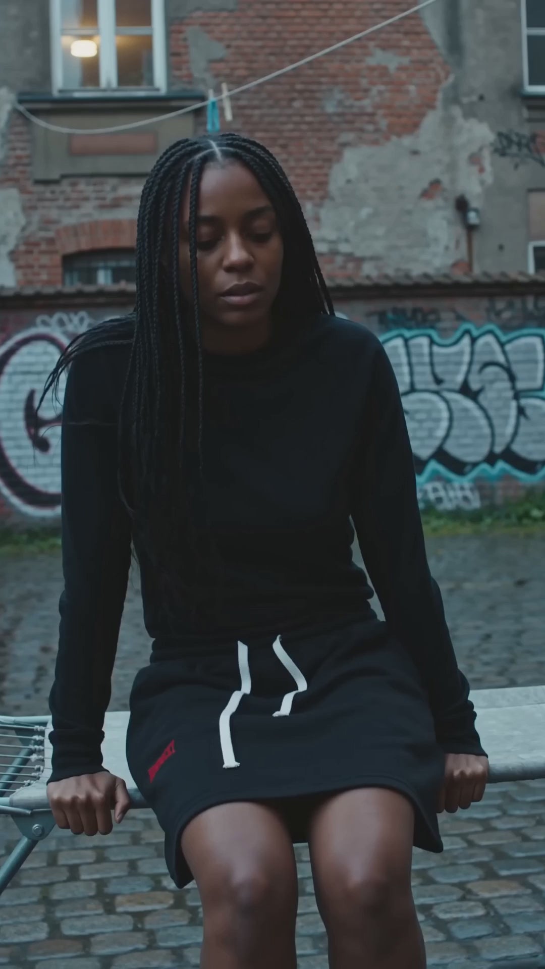 Spódnica Mini A-Line Nowrocky™ — kobieta z warkoczami siedzi na trzepaku na podwórku za kamienicą, czarna spódnica mini z białym nadrukiem, graffiti na ścianie.