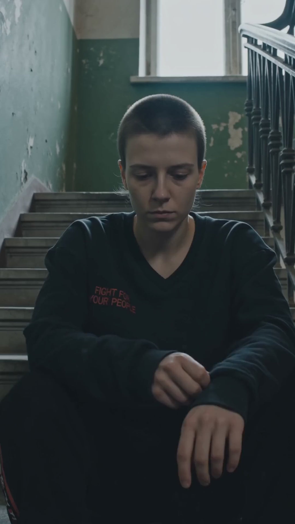Bluza Crop z Dekoltem V Nowrocky™ — osoba siedzi na schodach klatki schodowej kamienicy z opuszczoną głową, czarna krótka bluza z nadrukiem FIGHT FOR YOUR PEOPLE.