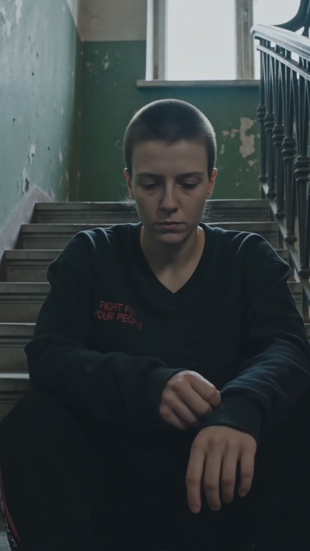 Bluza Crop z Dekoltem V Nowrocky™ — osoba siedzi na schodach klatki schodowej kamienicy z opuszczoną głową, czarna krótka bluza z nadrukiem FIGHT FOR YOUR PEOPLE.