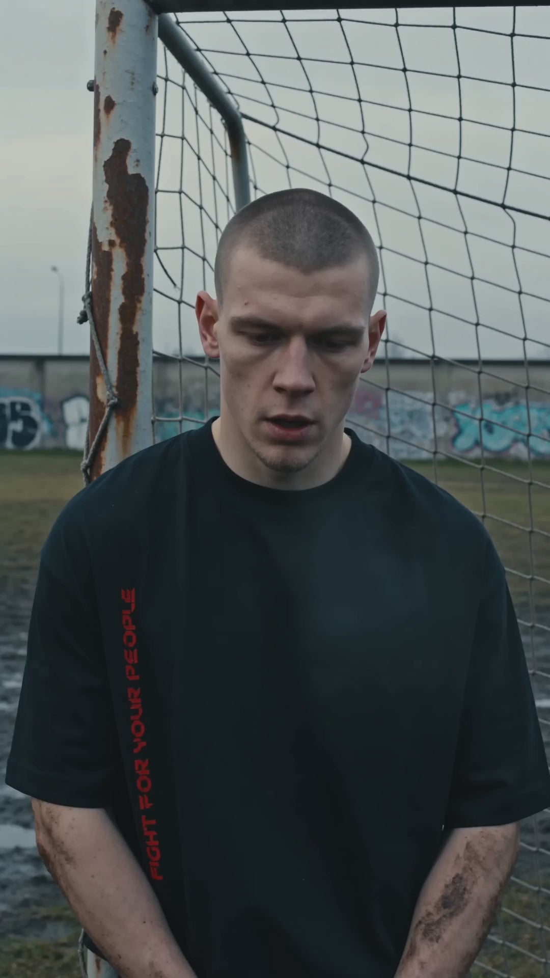 T-shirt Boxy Heavyweight Nowrocky™ — mężczyzna przed bramką na osiedlowym boisku, siatka za plecami, czarna koszulka z pionowym nadrukiem FIGHT FOR YOUR PEOPLE.