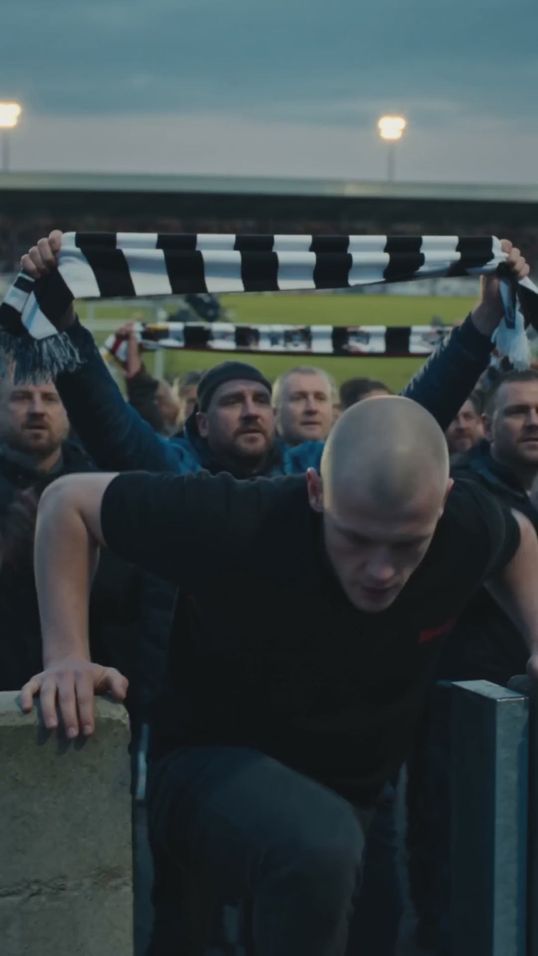 Polo Oversize Heavyweight Nowrocky™ — mężczyzna opiera się o barierkę na trybunie, za nim tłum trzyma nad głowami czarno-biały szalik, zmierzch.