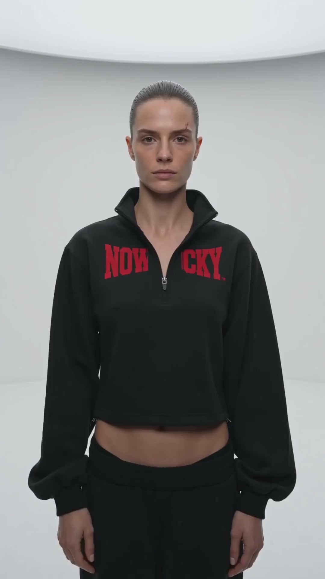 Bluza Crop Quarter-Zip ze Stójką Nowrocky™ — kobieta stoi po środku białego prześwietlonego studia, patrzy w obiektyw, czarna krótka bluza z czerwonym nadrukiem NOWROCKY na klatce.